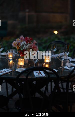 Delicati bicchieri da vino posti su un tavolo di legno scuro con illuminato candele e bouquet di fiori al centro di sera con bella fiori e alberi intorno Foto Stock