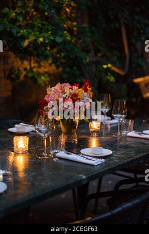 Delicati bicchieri da vino posti su un tavolo di legno scuro con illuminato candele e bouquet di fiori al centro di sera con bella fiori e alberi intorno Foto Stock