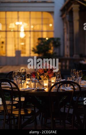 Delicati bicchieri da vino posti su un tavolo di legno scuro con illuminato candele e bouquet di fiori al centro di sera con bella fiori e alberi intorno Foto Stock