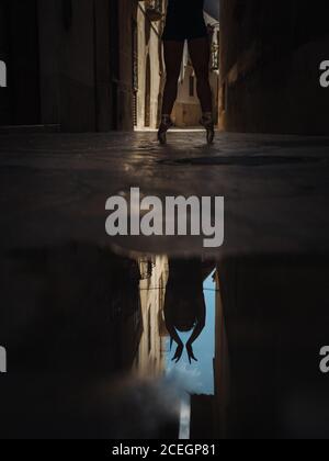 Crop ballerina di balletto irriconoscibile in piedi sulle dita dei piedi e riflettendo nel puddle sulla strada. Foto Stock