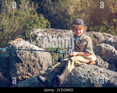 Ragazzo giovane con verga seduto e pesca su pietra grande in giornata di sole. Foto Stock