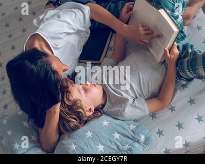 Dall'alto colpo di ragazzo dolce sdraiato su un bel letto e leggendo interessante libro con l'aiuto di madre. Foto Stock