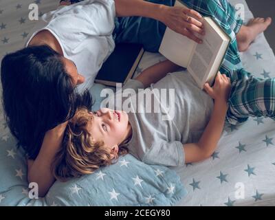 Dall'alto colpo di ragazzo dolce sdraiato su un bel letto e leggendo interessante libro con l'aiuto di madre. Foto Stock