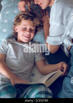 Dall'alto colpo di ragazzo dolce sdraiato su un bel letto e leggendo interessante libro con l'aiuto di madre. Foto Stock