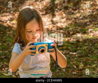 Piccola ragazza caucasica in una foresta che controlla la sua foto sopra la parte posteriore di una fotocamera digitale giocattolo in concentrazione Foto Stock