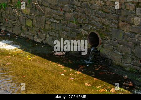 Un tubo di drenaggio in una parete di Cobblestone che versa in un Ruscello artificiale Foto Stock