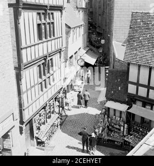 Francia, anni '50, vista storica dalla parte superiore di un vicolo stretto con negozi e open-air stand souvenir a le Mont-Saint-Michel, Normandia, un'isola marea e comune con l'antica abbazia benedettina. Il Monte St Michael in Cornovaglia, Regno Unito, anche se più piccolo, è simile a Saint-Michel, con un antico edificio costruito su un'isola di marea. Foto Stock