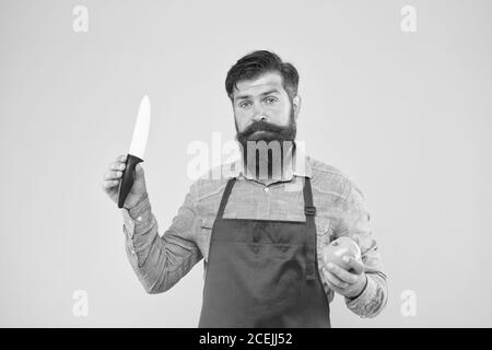 Cucina. Chef professionista in grembiule rosso. La cucina è il suo hobby. Uomo barbuto in uniforme cuoco. Casalinga maschio sicura preparare il pomodoro. Uomo maturo tenere coltello e verdure. Preparazione di insalata fresca. Foto Stock