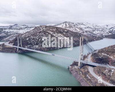 Pittoresca vista sul drone del lungo ponte che attraversa il bellissimo fiume tra magnifiche colline innevate nelle Asturie, Spagna Foto Stock