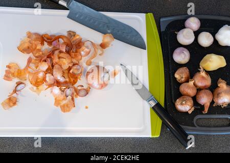 Cipolle di scalogno sbucciate con bucce di cipolla scartate su un bianco Tagliere in plastica con coltelli giapponesi Foto Stock