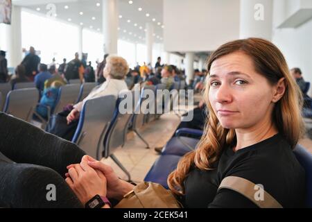 Giovane donna seduta nella sala dell'aeroporto, stanca dopo aver aspettato un paio d'ore per il volo di coincidenza la mattina presto, più passeggeri sfocati in ba Foto Stock