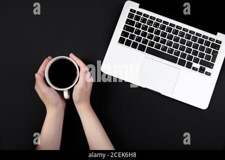 le mani tengono una tazza di caffè sul tavolo nero funzionante accanto al computer portatile Foto Stock