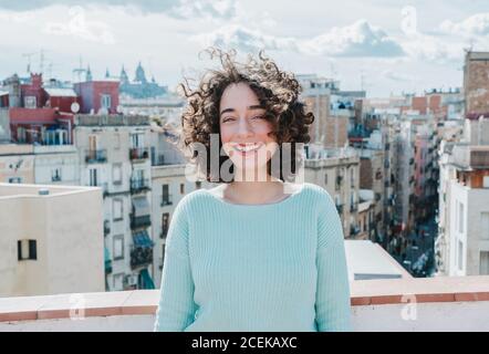 Bella giovane brunetta con capelli ricci che sorridono bianco in piedi sullo sfondo della bella città. Foto Stock