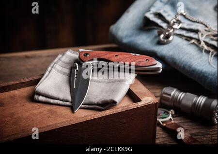 Coltello pieghevole in acciaio inox con manico in legno e altri articoli per il trasporto quotidiano (EDC) per uomo. Primo piano sul coltello pieghevole. Foto Stock