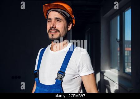 Un addetto alla costruzione regola un elmetto arancione sulla testa. Indossa una tuta da lavoro Foto Stock