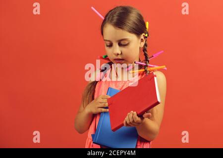 La ragazza tiene in mano libri rossi e blu. Torna a scuola e concetto d'arte. Pupilla in abito rosa con trecce piene di matite colorate e pennarelli. Ragazza scolastica con faccia interessata su sfondo rosso salmone Foto Stock