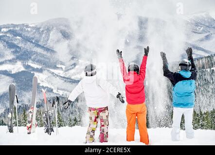 Vista posteriore di amici sciatori in tute da sci e caschi gettando neve fresca in alto in aria, divertendosi su una collina innevata con belle montagne sullo sfondo. Concetto di stazione sciistica e amicizia. Foto Stock