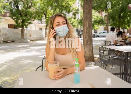 Giovane donna con maschera protettiva e disinfettante per le mani durante la videochiamata utilizzando il telefono cellulare nel bar della città. COVID-19 e New Normal, Health sa Foto Stock