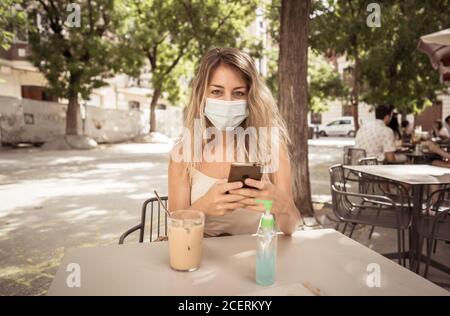 Giovane donna con maschera protettiva e disinfettante per le mani durante la videochiamata utilizzando il telefono cellulare nel bar della città. COVID-19 e New Normal, Health sa Foto Stock