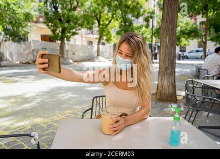 Giovane donna con maschera protettiva e disinfettante per le mani durante la videochiamata utilizzando il telefono cellulare nel bar della città. COVID-19 e New Normal, Health sa Foto Stock