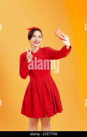 Giovane felice attraente femmina in abito rosso scatta foto utilizzando il suo telefono cellulare, sparare su sfondo arancione colorato Foto Stock