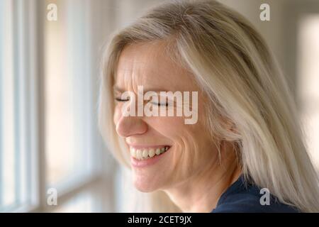 Felice bionda di mezza età che sogna con un sorriso luminoso di piacere come lei si appoggia su un davanzale che guarda all'esterno dentro primo piano Foto Stock