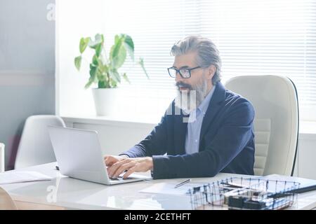 Ritratto di un operaio maschile concentrato che indossa occhiali seduti alla scrivania dell'ufficio che scriva qualcosa sul computer portatile Foto Stock