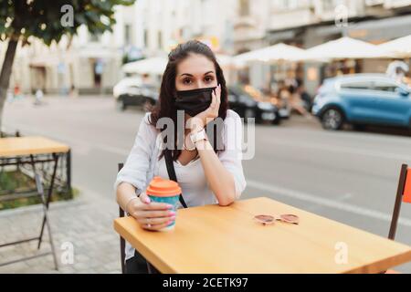 Giovane donna seduta sulla strada in una maschera con una tazza di caffè. Foto Stock