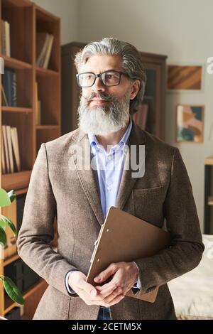 Uomo moderno e maturo con bearded e outfit elegante che tiene gli appunti guardando lontano, verticale medio ritratto Foto Stock