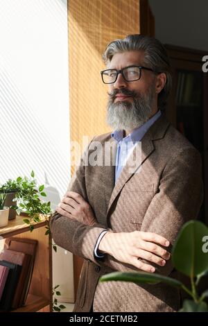 Uomo dal braccio maturo sicuro che indossa un abito elegante e occhiali in piedi con le braccia incrociate guardando la fotocamera, verticale medio ritratto Foto Stock