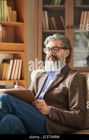 Immagine verticale media di un bell'uomo con bearded che indossa un abito alla moda seduta sul divano in una moderna sala da ufficio per prendere appunti Foto Stock