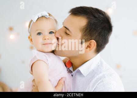 Il giovane papà felice tiene una figlia sorridente di un anno nella sua braccia e la bacia sulla guancia dei bambini camera Foto Stock