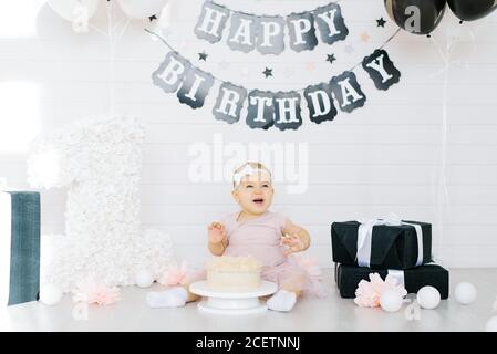 Una ragazza di un anno si siede sul pavimento vicino ad una dolce torta in onore del suo primo compleanno, prova la torta e sorride, ride Foto Stock