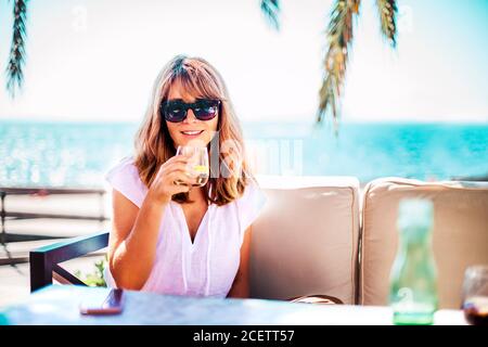 Foto ritratto di donna felice godendo del sole estivo e bevendo un bicchiere d'acqua mentre si rilassa sul divano del salotto vicino al mare. Foto Stock