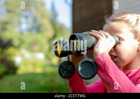 Bambina bionda che guarda attraverso il binocolo sullo sfondo della natura. Il bambino si nascose nel verde e si siede in agguato. Concetto di avventura per bambini. Il bambino Foto Stock