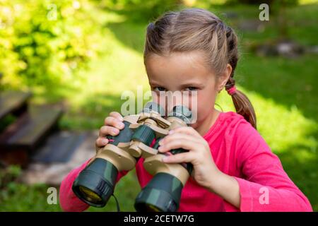 Bambina bionda che guarda attraverso il binocolo sullo sfondo della natura. Il bambino si nascose nel verde e si siede in agguato. Concetto di avventura per bambini. Il bambino Foto Stock