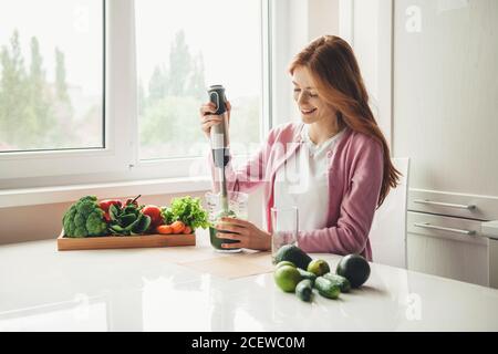 Signora zenzero con frettole utilizzando uno spremiagrumi elettrico fare fresco succo in cucina di lime e avocado Foto Stock