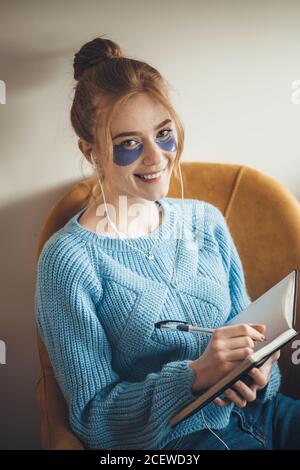 Primo piano foto di una donna caucasica con macchie di occhio e frettoliamo sorridendo alla fotocamera mentre si ascolta la musica e. scrivere qualcosa nel libro Foto Stock