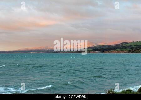 Montagne illuminate al tramonto e città in lontananza sul Mare d'Irlanda a Trefor, Galles del Nord Foto Stock
