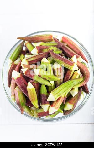 Okra verde e rosso, pulito e preparato per cucinare in ciotola su fondo di legno. Il dito della donna nella ciotola. Foto Stock