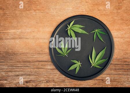 Foglie di cannabis, mariuana di diverse dimensioni su piatto nero e tavolo di legno Foto Stock