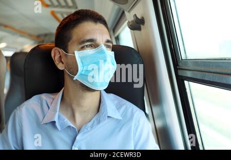 Viaggiare in sicurezza con i mezzi pubblici. Giovane uomo con maschera chirurgica che guarda attraverso la finestra del treno. Addestrare il passeggero con una maschera protettiva che si affaccia attraverso Foto Stock
