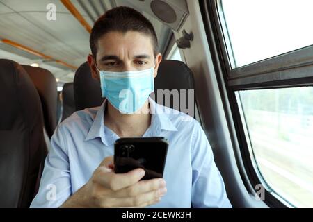Uomo con maschera chirurgica utilizzando l'app per smartphone sui mezzi pubblici. Addestrare il passeggero con maschera protettiva che viaggia seduto in business class testando sopra Foto Stock