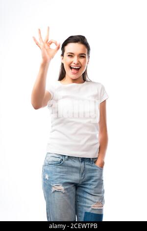 giovane donna in t-shirt bianca e jeans in piedi con la mano nello slot e con il segno ok isolato su bianco Foto Stock