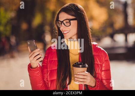 Foto di bella moda ragazza riposo relax weekend autunno parco aspetta un amico usa lo smartphone leggi sms hold take out cappuccino beverage tazza indossare un cappotto rosso Foto Stock