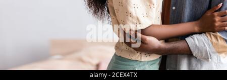 vista ritagliata della coppia afroamericana in abiti casual abbracciando mentre in piedi a casa, intestazione del sito web Foto Stock