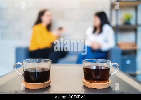 Fuoco selettivo di coppia di tazze di vetro di nero caldo caffè messo sul tavolo in un ambiente di lavoro contemporaneo con donna sfocata colleghi che parlano in background Foto Stock