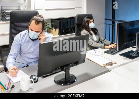 Serio giovane uomo messo a fuoco in maschera medica che discute di problema di affari per telefono mentre una collega femminile lavora con il computer in contemporanea ufficio di codorking Foto Stock