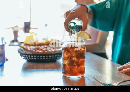 Coltivare il maschio irriconoscibile versando la bevanda fredda dalla bottiglia in vetro tazza con ghiaccio e fetta di limone mentre si pranza con amici al caffè Foto Stock