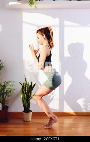 Vista laterale della donna serena in Utkatasana pratica la consapevolezza mentre fare yoga in una stanza accogliente a casa Foto Stock